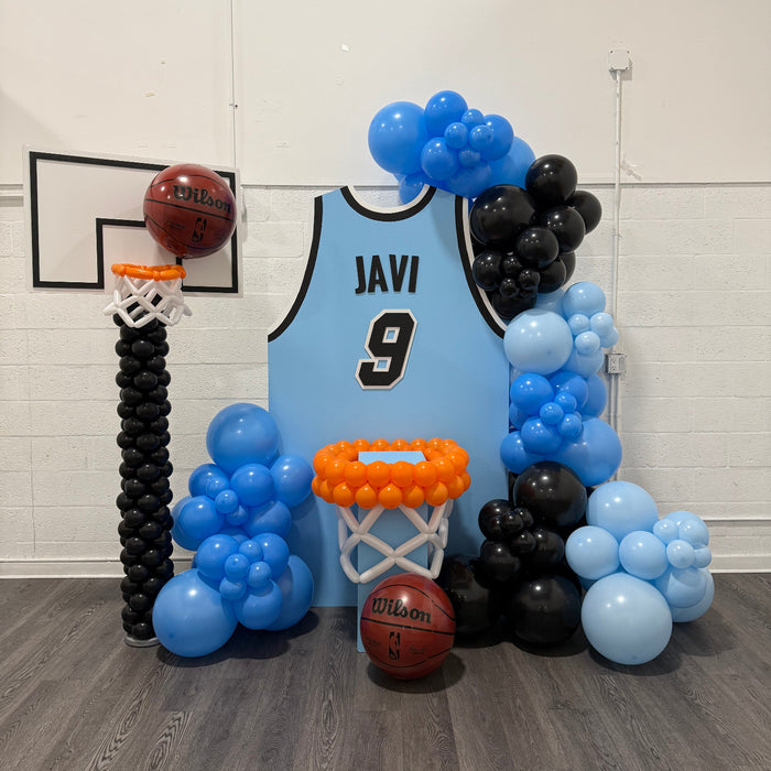 Basketball Jersey Backdrop featuring a custom light blue jersey with black trim, personalized name, and number centerpiece surrounded by blue and black balloon garlands. Complete with balloon basketball hoops and real Wilson basketballs, this setup is perfect for pep rallies, senior nights, team banquets, and basketball-themed events. Designed using the Basketball Jersey Backdrop Template from The Creative Heart Studio.