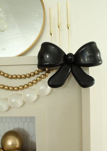 Close-up of an elegant black balloon bow styled on a white mantel, created by Lilly Jimenez. Accented with gold pearl balloon garland and taper candles, this classy balloon decor is perfect for New Year’s Eve parties, weddings, or sophisticated celebrations. A stylish detail from the Champagne Balloon Bottle & Big Balloon Bow Tutorial, ideal for upscale balloon installations and cocktail hour decor.