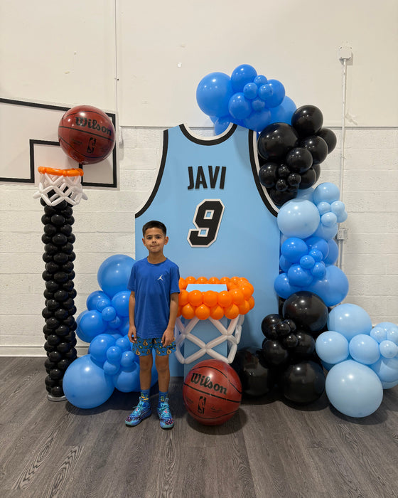 A young boy stands in front of a custom Basketball Jersey Backdrop featuring a light blue jersey with name and number, surrounded by black and blue balloon garlands, basketball hoops, and Wilson basketball props. Perfect for pep rallies, team banquets, and sports-themed parties, this backdrop captures the excitement of game day. Designed using the Basketball Jersey Backdrop Template from The Creative Heart Studio.