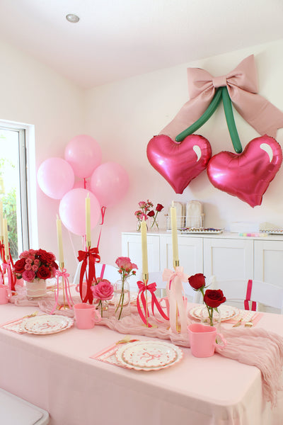 Elegant pink Galentine’s Day party setup featuring a cherry-inspired balloon backdrop with heart-shaped balloons, a large bow, and green balloon stems. A beautifully decorated table includes pink tableware, red roses, tall candles with ribbon accents, and soft pink linens, creating a dreamy and festive celebration atmosphere.