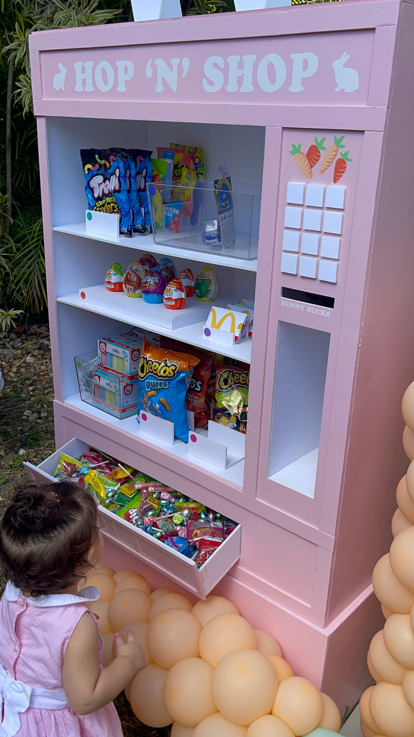 A pastel pink foamboard vending machine prop labeled “Hop ‘N’ Shop” is filled with candy, chips, and snacks. A young child explores the pull-out drawer of treats, adding charm to this Easter or kids’ party setup. The vending machine features bunny and carrot details, foamboard construction, and balloon decor. This prop is taught in the Foamboard Vending Machine Class and is perfect for photo ops and candy-themed events.