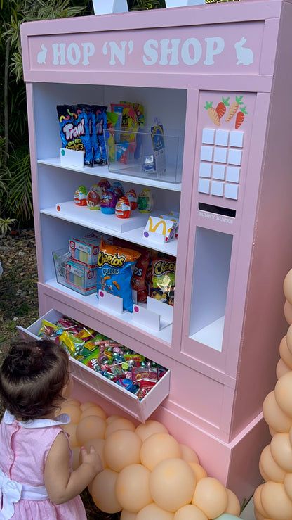A pastel pink foamboard vending machine prop labeled “Hop ‘N’ Shop” is filled with candy, chips, and snacks. A young child explores the pull-out drawer of treats, adding charm to this Easter or kids’ party setup. The vending machine features bunny and carrot details, foamboard construction, and balloon decor. This prop is taught in the Foamboard Vending Machine Class and is perfect for photo ops and candy-themed events.