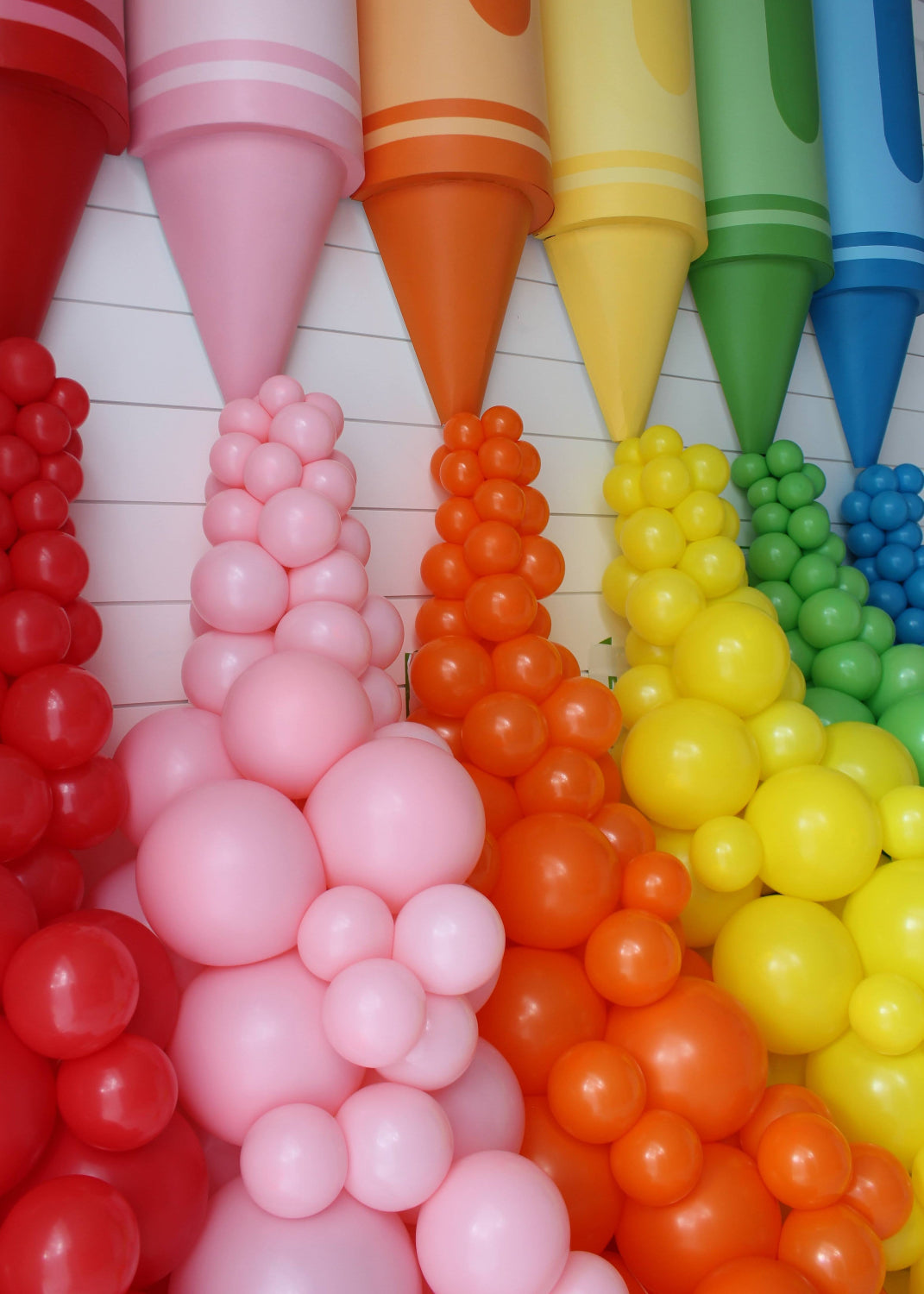 Zoomed-in view of oversized crayon props in red, pink, orange, yellow, green, and blue with coordinating balloon garlands cascading from each crayon tip. This colorful and playful classroom or party display was created by Lilly Jimenez as part of her Crayon Prop Tutorial for Creative Heart Studio, perfect for back-to-school setups, teacher appreciation, or creative photo backdrops.