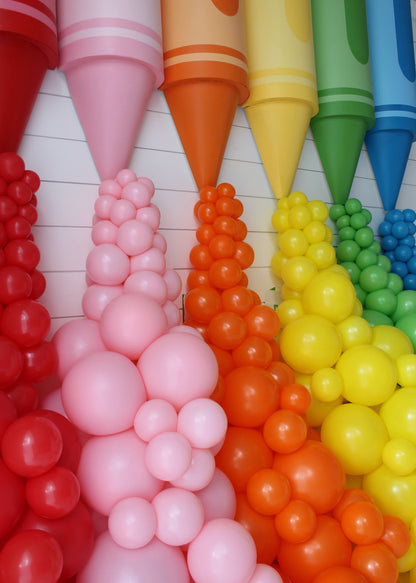 Zoomed-in view of oversized crayon props in red, pink, orange, yellow, green, and blue with coordinating balloon garlands cascading from each crayon tip. This colorful and playful classroom or party display was created by Lilly Jimenez as part of her Crayon Prop Tutorial for Creative Heart Studio, perfect for back-to-school setups, teacher appreciation, or creative photo backdrops.