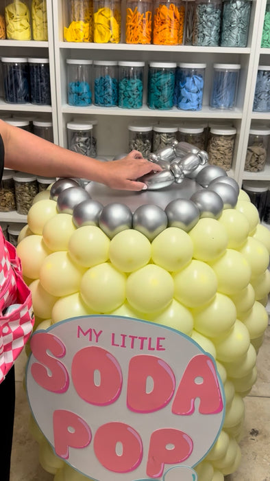 Close-up of a K-pop Demon Hunters inspired balloon soda pop prop featuring pastel yellow balloons forming the can, a silver balloon tab on top, and a “My Little Soda Pop” label, shown in a studio with organized balloon supplies in the background.