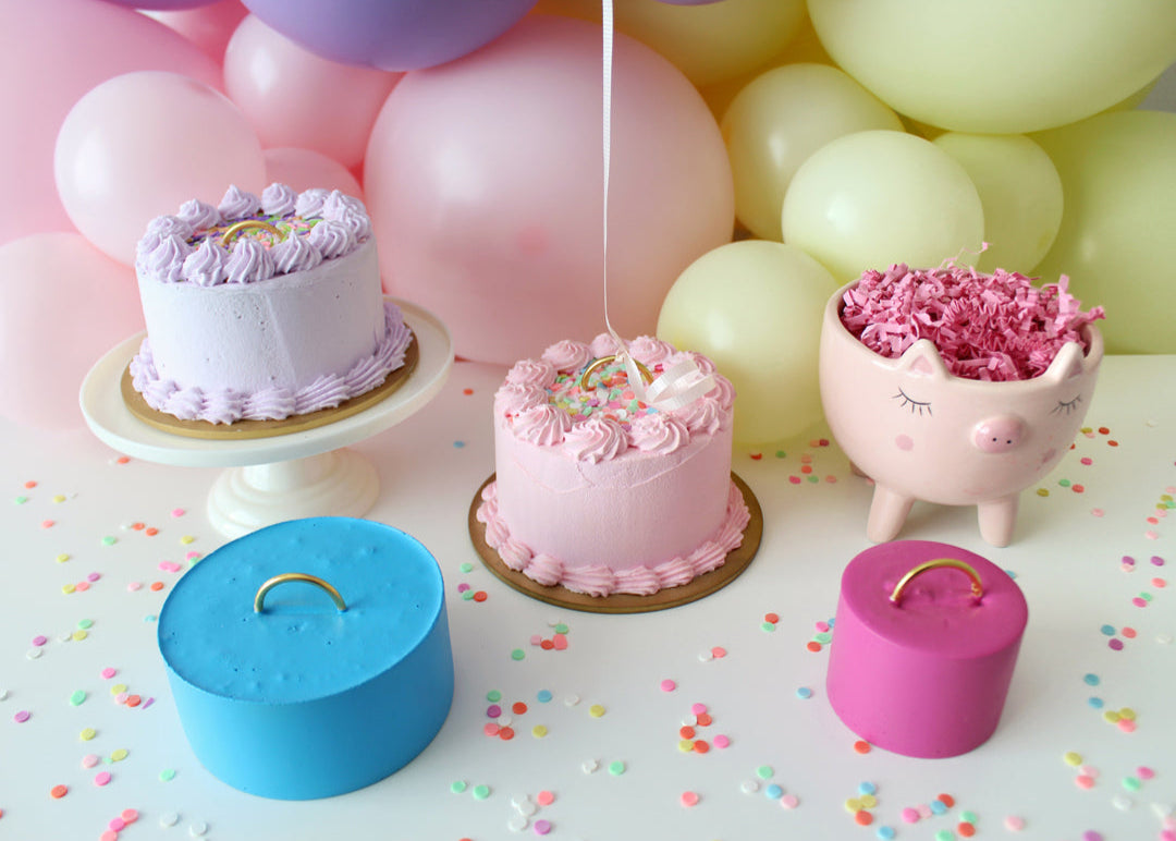 A pastel balloon styling scene featuring faux cake balloon weights and colorful DIY weights with gold handles. Designed by Lilly Jimenez, the pink and purple cakes sit among festive balloons, confetti, and a cute pig planter. This setup is from Lilly’s Balloon Weights Class, where she teaches you to make three adorable styles—plus a bonus trick using a simple household item for a creative and cost-saving weight solution.