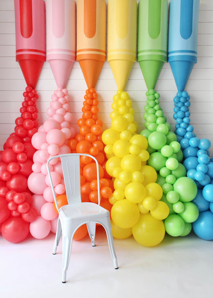 Oversized rainbow crayon props with matching balloon garlands displayed against a white backdrop, featuring a white chair placed in front to show the impressive scale of the crayon designs. Created by Lilly Jimenez of Creative Heart Studio, this colorful setup is part of the Crayon Prop Tutorial and ideal for back-to-school parties, classroom decor, or playful photo booth installations.