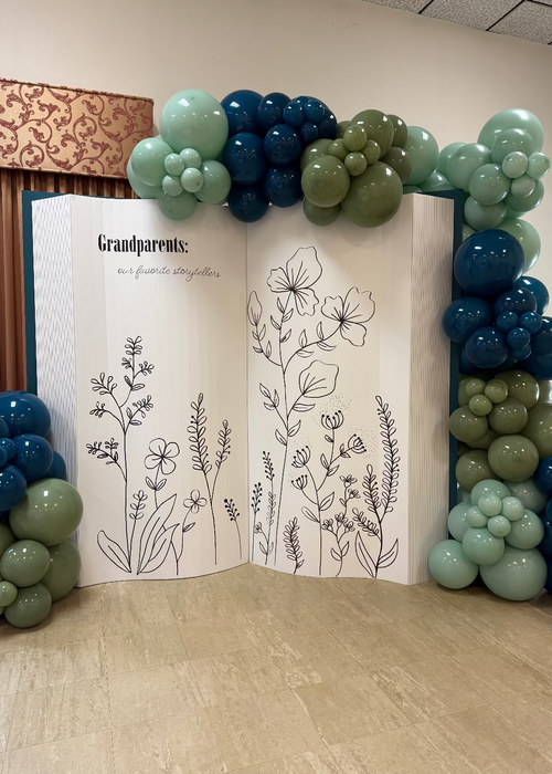 DIY Open Book Backdrop decorated with clusters of green and blue balloons, featuring floral illustrations inside the oversized foamboard book. This creative backdrop is perfect for storybook themes, graduations, weddings, and photo booths. Built using a digital download template, it’s a customizable project for event decorators and balloon stylists looking to add a unique statement piece.