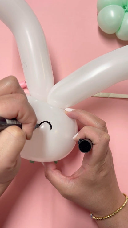Hands drawing a black eye with a marker on a white bunny balloon head with long ears. This behind-the-scenes view shows a key step in creating a balloon bunny topper, perfect for Easter candy cups, party favors, or balloon art tutorials. Pink background and mint green balloon parts complete the cheerful look.