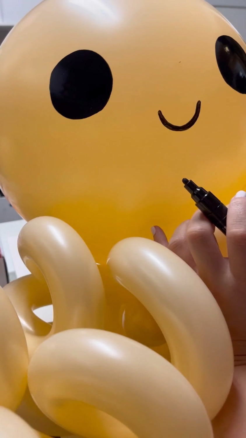 Close-up of a hand drawing a cute smile on a balloon octopus using a black marker. This balloon octopus is part of the Under the Sea Balloon Class, perfect for ocean-themed decorations and kids' party designs.
