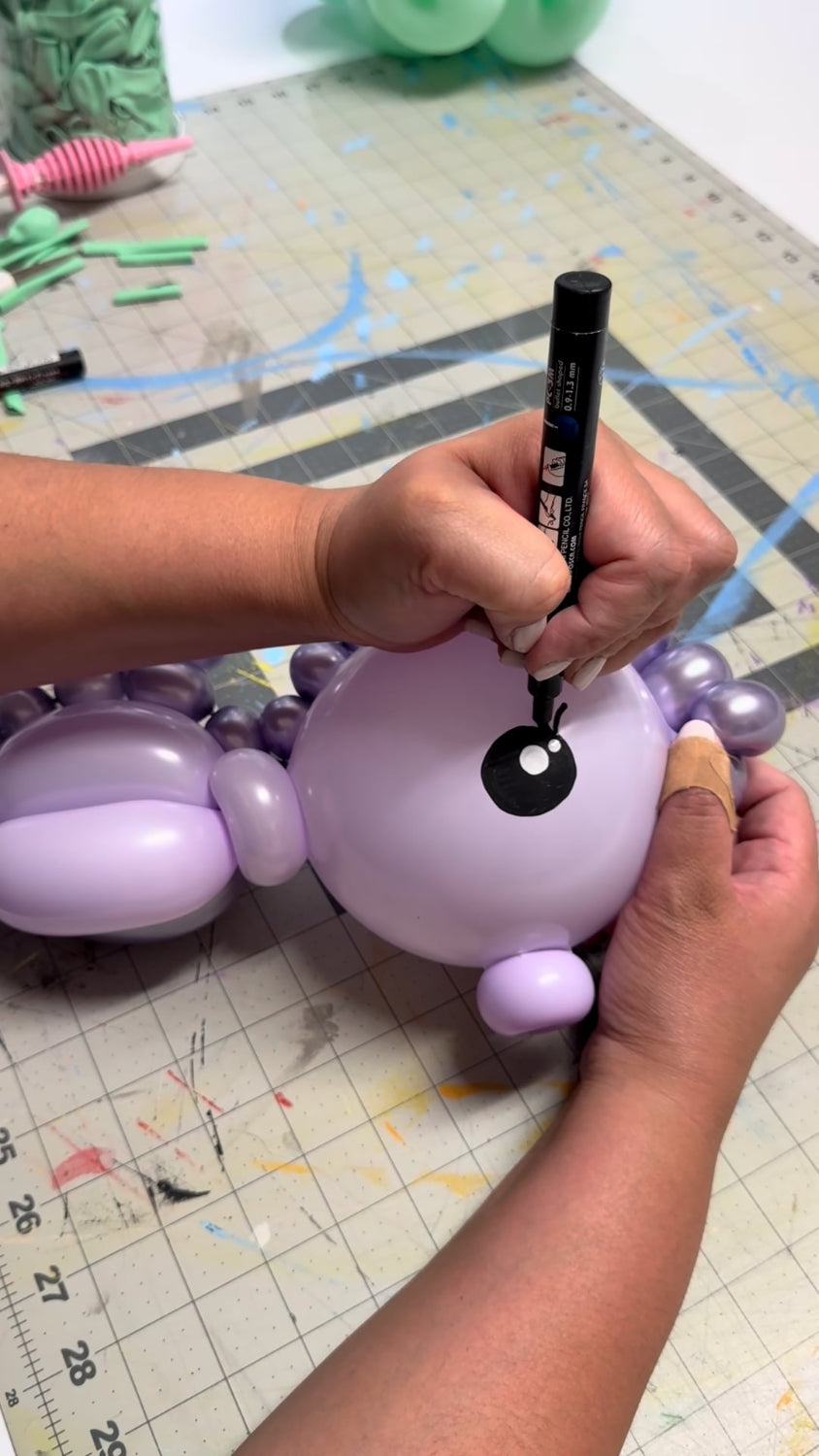 Close-up of a hand drawing an eye with a black paint pen onto a lavender balloon seahorse during the Under the Sea Balloon Class by Lilly Jimenez. The balloon sculpture is positioned on a craft mat with tools and balloons visible in the background. This step is part of the finishing touches shown in the class’s step-by-step tutorial, teaching decorators how to add personality and detail to their balloon sea creatures.