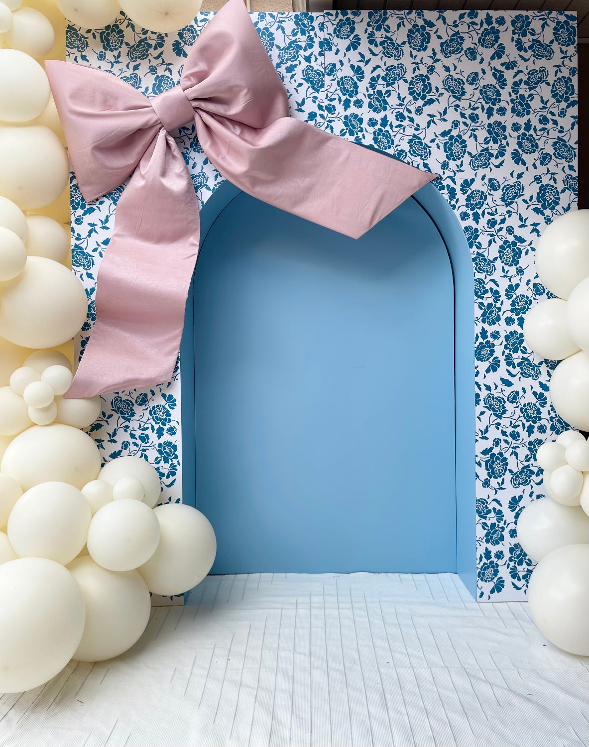 A beautifully styled event setup featuring a blue arched doorway framed by a floral-patterned backdrop. An oversized dusty pink bow is placed at the top left corner, adding a soft, elegant touch. Cream-colored balloon clusters surround the arch, creating a dreamy and sophisticated atmosphere perfect for special occasions.