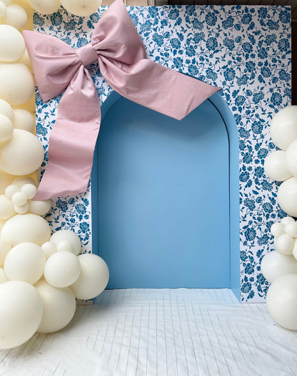 A beautifully styled event setup featuring a blue arched doorway framed by a floral-patterned backdrop. An oversized dusty pink bow is placed at the top left corner, adding a soft, elegant touch. Cream-colored balloon clusters surround the arch, creating a dreamy and sophisticated atmosphere perfect for special occasions.
