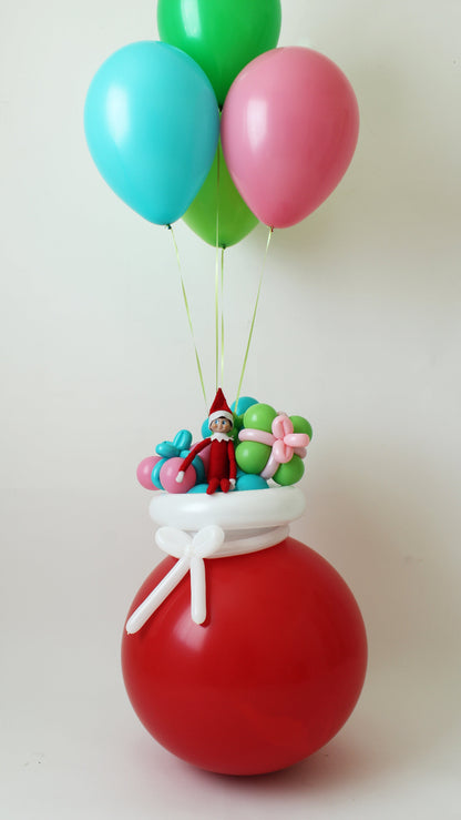 Whimsical Elf Arrival balloon Santa sack created by Lilly Jimenez, featuring a red balloon base tied with a white balloon bow. On top sits an Elf on the Shelf surrounded by pastel balloon presents, with three helium balloons in pink, green, and teal floating above. A magical and festive balloon decor idea for Christmas or Elf on the Shelf return displays.