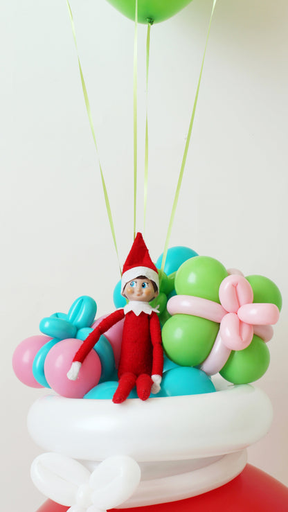 Close-up of Elf on the Shelf sitting atop a white balloon sack lid, surrounded by pastel balloon presents in pink, green, teal, and blue. Part of Lilly Jimenez’s Elf Arrival Santa Sack tutorial, this creative balloon design makes the perfect whimsical touch for Elf arrival displays, holiday decor, or Christmas morning surprises.
