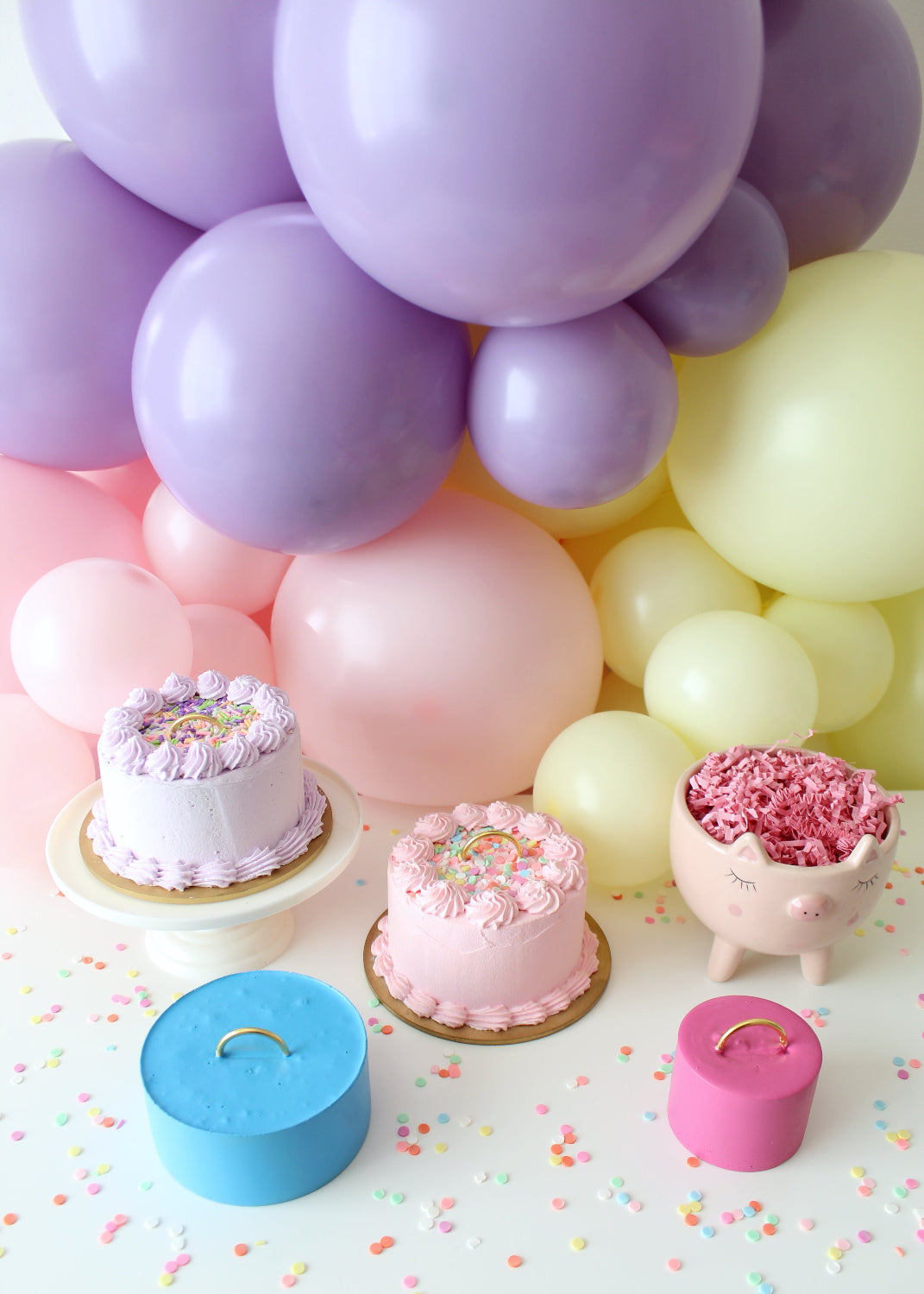 Pastel party scene with faux cake balloon weights by Lilly Jimenez. Two cake-shaped weights with piped icing and colorful sprinkles sit among a balloon garland of purples, pinks, and yellows. A pig planter and DIY balloon weights with gold handles complete the whimsical setup from Lilly’s Balloon Weights Class—where you'll learn to make three styles and discover a clever, cost-effective bonus method.