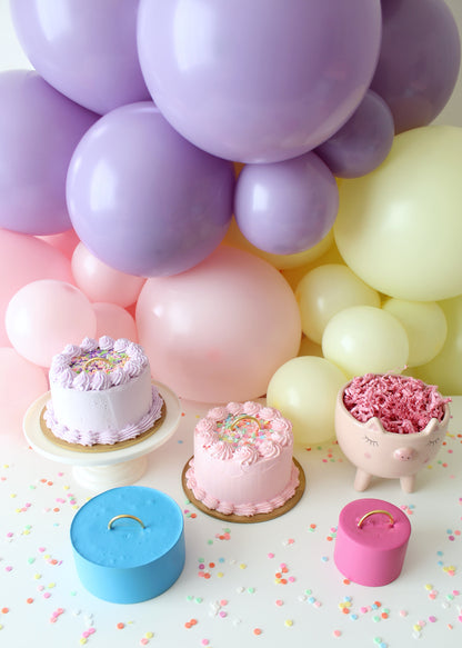Pastel party scene with faux cake balloon weights by Lilly Jimenez. Two cake-shaped weights with piped icing and colorful sprinkles sit among a balloon garland of purples, pinks, and yellows. A pig planter and DIY balloon weights with gold handles complete the whimsical setup from Lilly’s Balloon Weights Class—where you'll learn to make three styles and discover a clever, cost-effective bonus method.