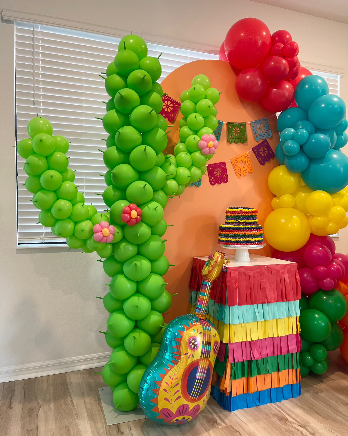 Close-up of a vibrant balloon cactus column decorated with mini balloon flowers, part of a colorful fiesta-themed party setup. Includes a rainbow fringe-covered table with a striped cake topper, papel picado banner, and a fiesta guitar balloon prop—ideal for Cinco de Mayo or cactus birthday décor.