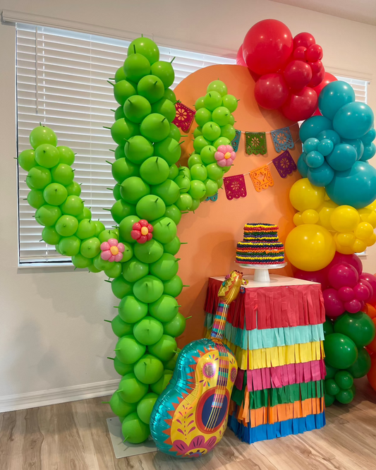 Close-up of a vibrant balloon cactus column decorated with mini balloon flowers, part of a colorful fiesta-themed party setup. Includes a rainbow fringe-covered table with a striped cake topper, papel picado banner, and a fiesta guitar balloon prop—ideal for Cinco de Mayo or cactus birthday décor.