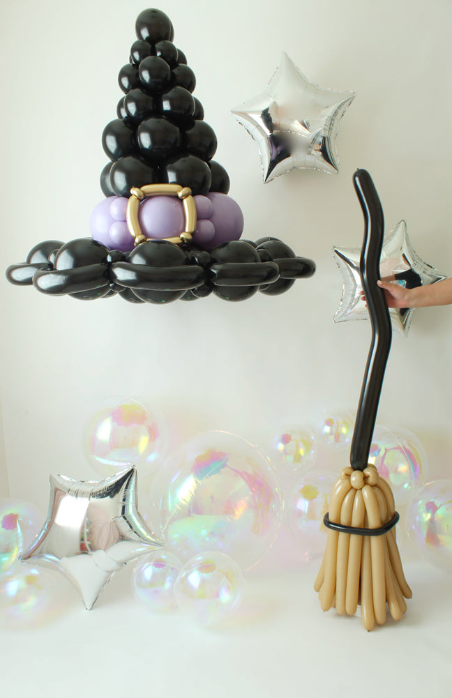 A floating black balloon witch hat with a gold buckle and purple accents hovers above a twisted balloon broomstick, creating a whimsical and magical Halloween display. Silver star balloons and iridescent bubbles add to the enchanting atmosphere. This design is perfect for Halloween parties, themed events, and festive photo backdrops. Learn to make these spooky balloon props in the Witch Hat & Broom Balloon Tutorial!