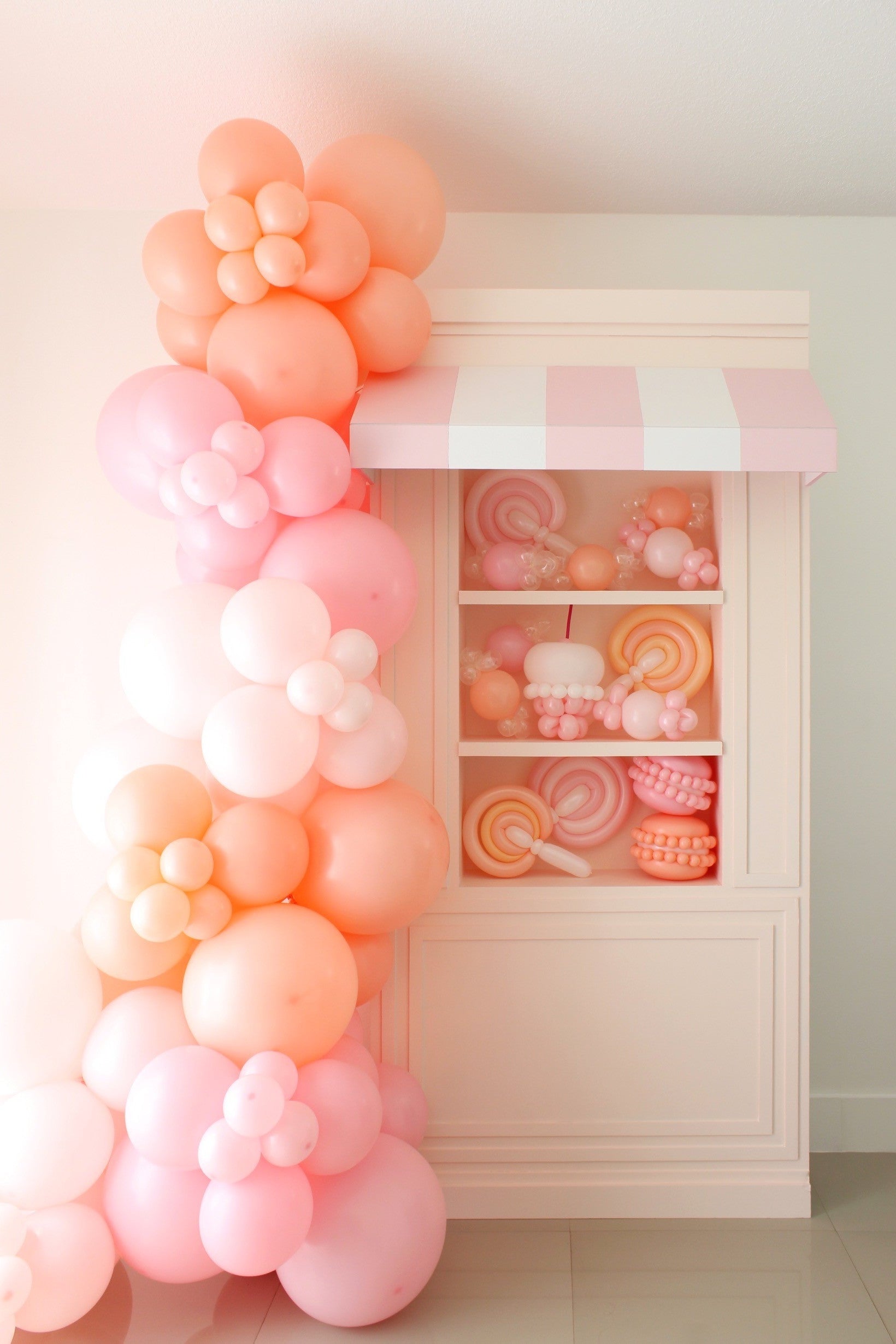 DIY foamboard storefront display featuring a pink-and-white striped awning, built-in shelving filled with pastel balloon candy props like lollipops and macarons, and a large cascading garland made of peach and pink balloons shaped into flowers. Part of the Foamboard Storefront Class from The Creative Heart Studio—perfect for birthday parties, candy themes, and event decor.