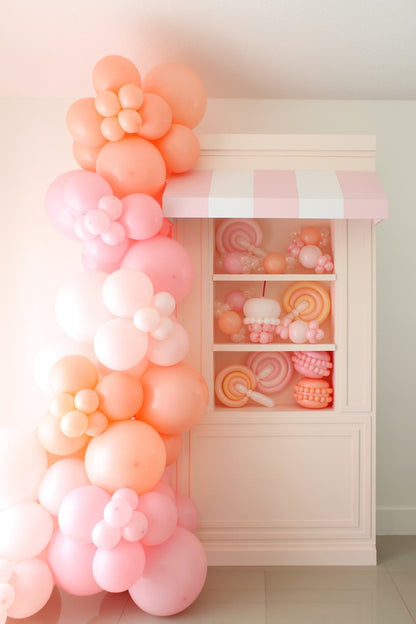 DIY foamboard storefront display featuring a pink-and-white striped awning, built-in shelving filled with pastel balloon candy props like lollipops and macarons, and a large cascading garland made of peach and pink balloons shaped into flowers. Part of the Foamboard Storefront Class from The Creative Heart Studio—perfect for birthday parties, candy themes, and event decor.