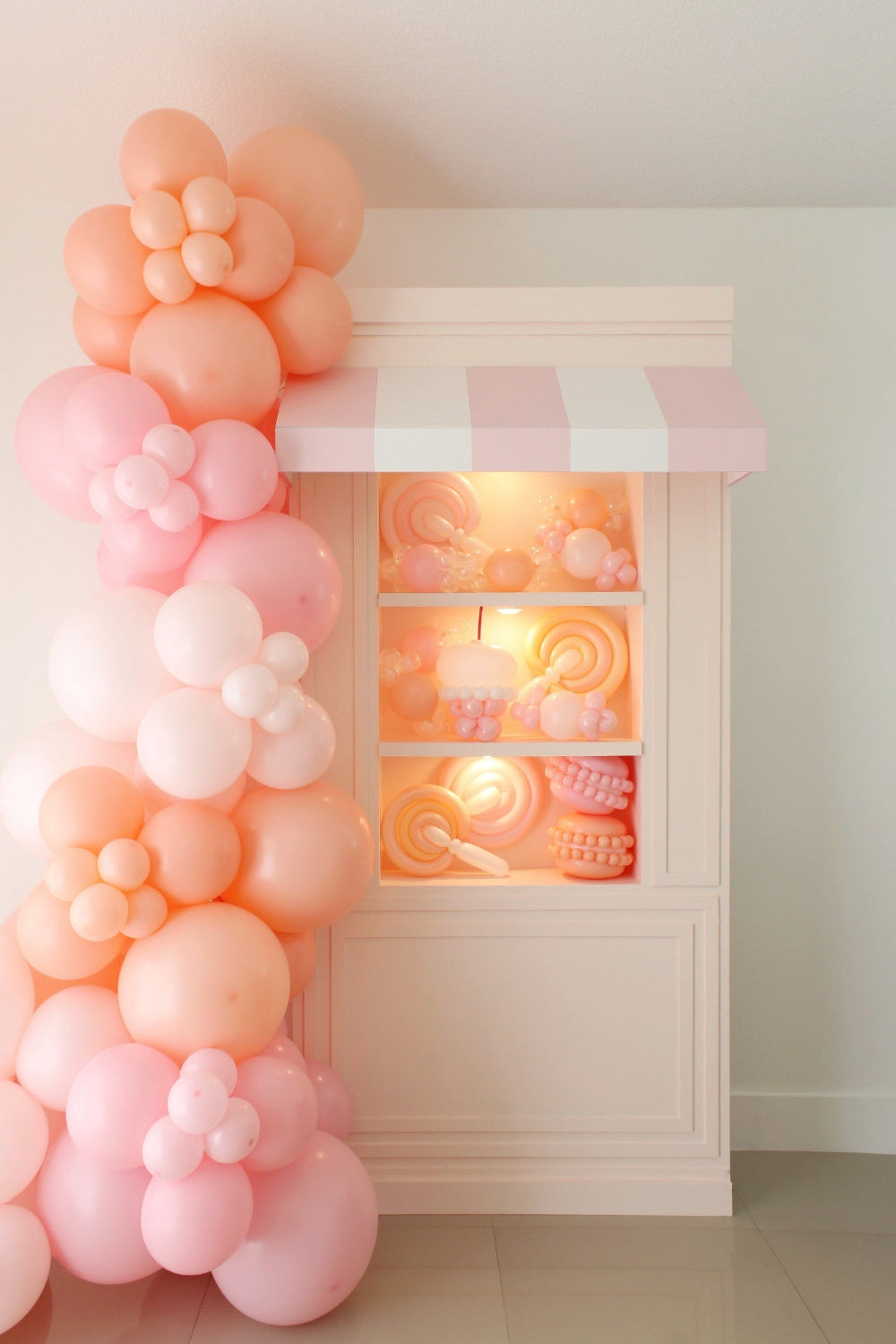 Full view of a life-size foamboard storefront prop featuring a pink-and-white striped awning, built-in shelves filled with pastel balloon candy creations, and a cascading balloon garland in peach and pink tones. A perfect DIY setup for birthdays, candy-themed parties, and photo ops—part of the Foamboard Storefront Class from The Creative Heart Studio.