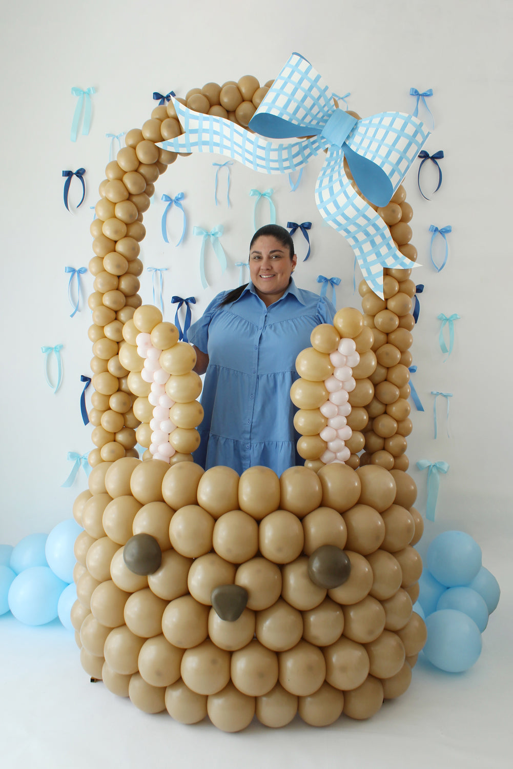 Lilly stands inside a giant balloon Easter bunny basket made from brown balloons, complete with a blue gingham bow and bunny face details. This adorable creation is part of the Easter Balloon Class and makes a perfect photo op or centerpiece for spring events and Easter celebrations.