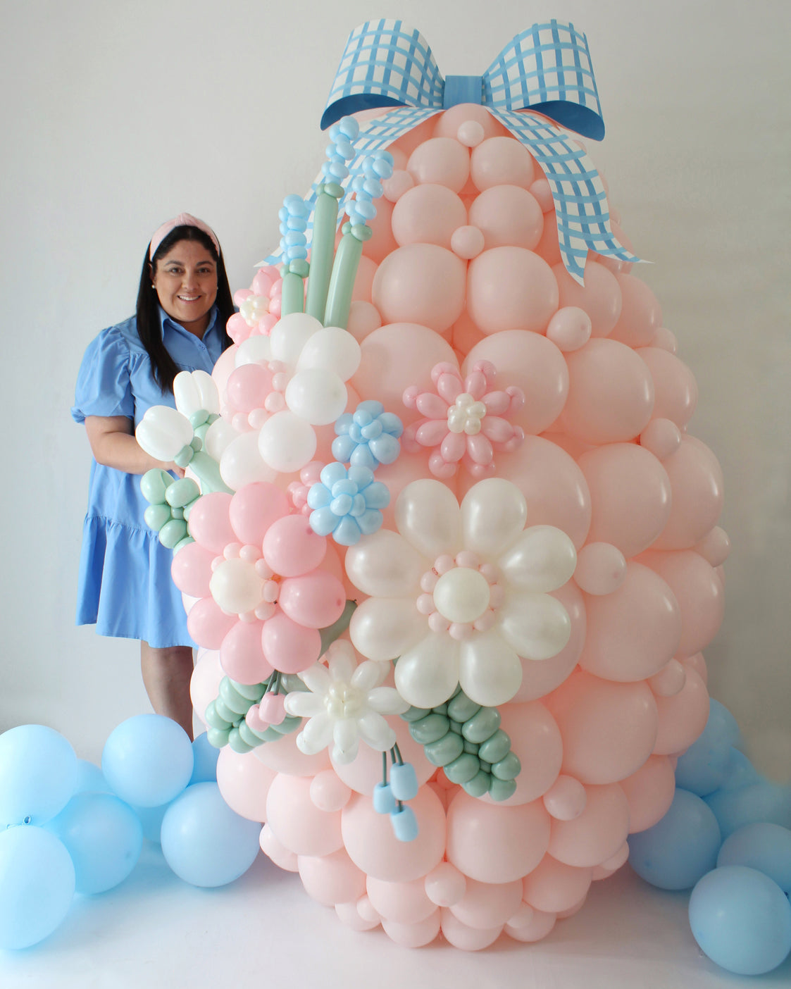 Lilly poses next to a giant pastel pink balloon Easter egg covered in 3D balloon flowers and topped with a blue gingham bow. A show-stopping design from her Easter Balloon Class, perfect for spring-themed backdrops and Easter celebrations.