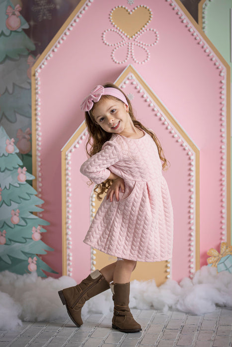Little girl wearing a pink quilted dress, matching bow headband, and brown boots posing confidently in front of a pastel pink gingerbread house backdrop with candy details, faux snow, and Christmas trees—perfect for holiday photo sessions and festive decor inspiration.