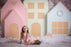 Little girl wearing a pink dress and bow posing in front of an oversized pastel gingerbread village backdrop, featuring pink, mint, and blue foam board houses with candy-style trim, pearl wreaths, faux snow, and string lights—ideal for holiday photo sessions and Christmas party decor.