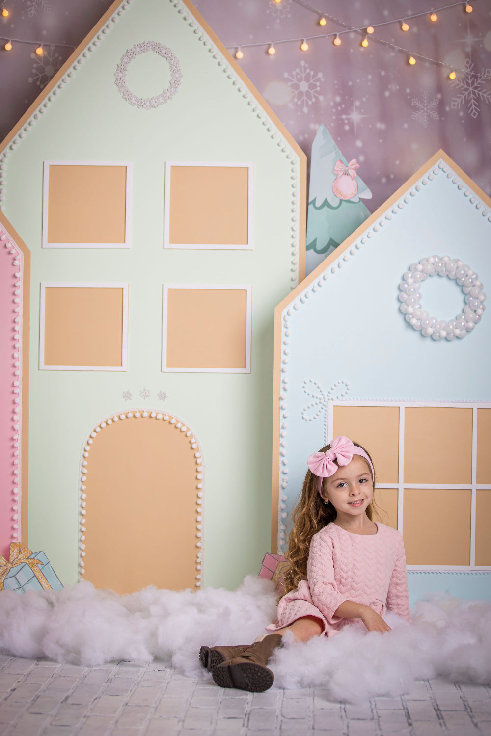 Little girl in a pink dress and matching bow sitting on fluffy faux snow in front of a pastel gingerbread village backdrop, featuring oversized pink, mint, and blue houses decorated with candy details and holiday string lights—perfect for Christmas photo sessions and festive party decor.