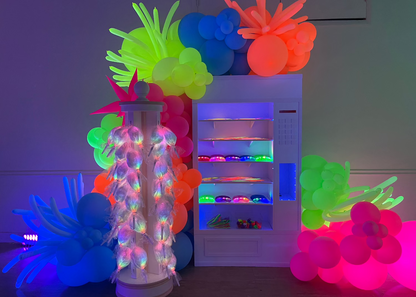 This foamboard vending machine is styled with glow-in-the-dark and LED elements, surrounded by vibrant neon balloon garlands. The display includes LED-lit cotton candy and colorful light-up party favors, perfect for a glow party or 80s theme. Built using the Foamboard Vending Machine Class tutorial, this prop is ideal for party planners looking to create a bold, interactive photo backdrop or snack station.