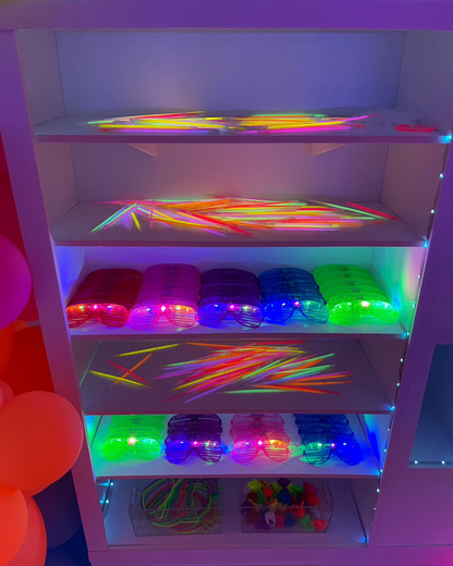 Close-up of foamboard vending machine shelves styled for a glow party. Each shelf is stocked with neon glow sticks, LED glasses, light-up accessories, and vibrant party favors under UV lighting. The glowing items are arranged in rainbow order for visual impact. This colorful vending machine setup is featured in the Foamboard Vending Machine Class and is ideal for glow-in-the-dark or 80s-themed celebrations.