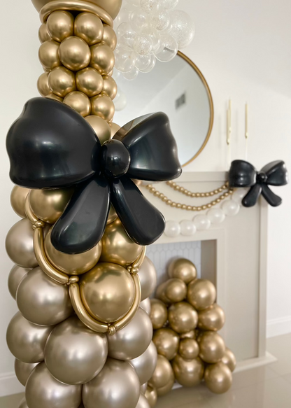 Elegant gold and pearl balloon champagne bottle with a large black balloon bow, styled by Lilly Jimenez. The display includes a matching balloon bow on the fireplace and gold balloon accents, perfect for weddings, New Year's Eve, and upscale parties. A stunning photo op and focal point for any celebration.