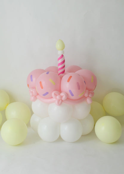 Whimsical balloon cake featuring pastel pink balloon frosting with sprinkle details, white balloon base, and a striped candle topper. Surrounded by pale yellow balloons, this balloon sculpture is ideal for birthday decorations, party setups, and balloon art classes. A sweet and fun design that adds charm to any celebration.