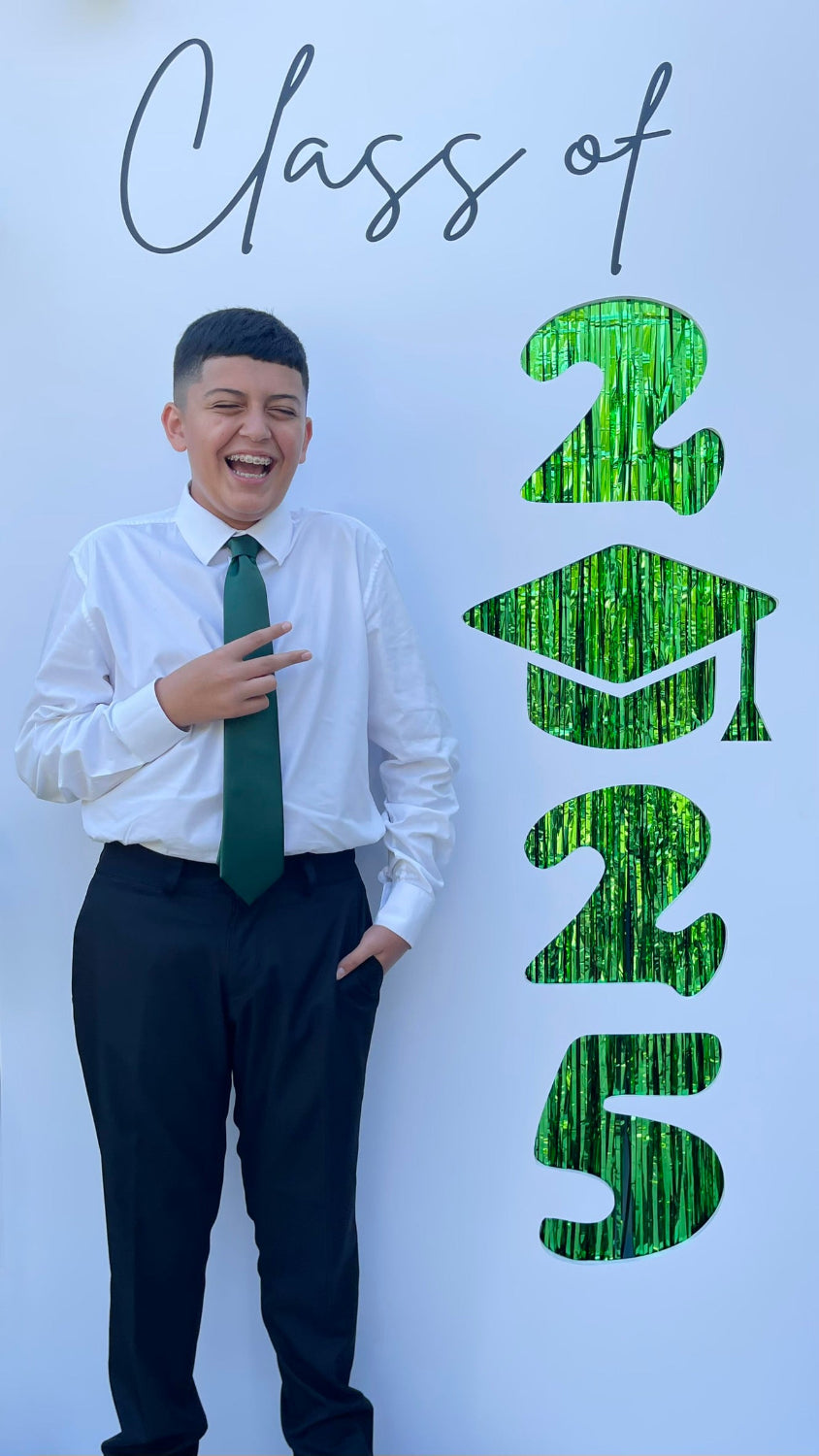 Smiling student in dress shirt and green tie posing next to a tall “Class of 2025” graduation photo backdrop. The numbers are cut out vertically with shiny green fringe in the background and a graduation cap replacing the zero. Perfect for preschool, middle school, high school, or college graduation photo ops.