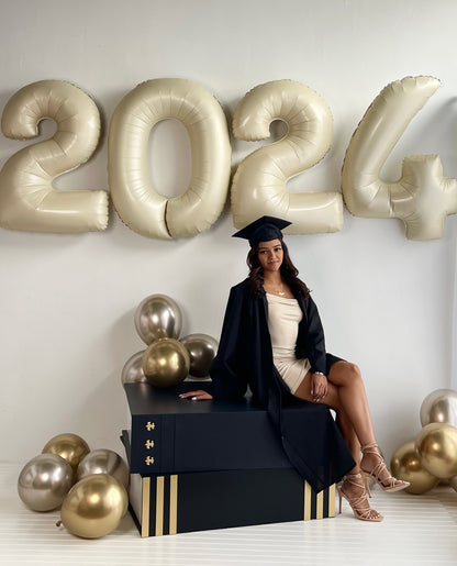A graduate in a cap and gown sits elegantly on a black and gold oversized book prop, surrounded by gold and champagne balloons. Behind her, large gold 2024 balloons create a celebratory backdrop. This setup, designed by Lilly Jimenez of The Creative Heart Studio, is perfect for graduation photoshoots, event decor, and literary-themed celebrations. Part of the Oversized Book Prop Class!