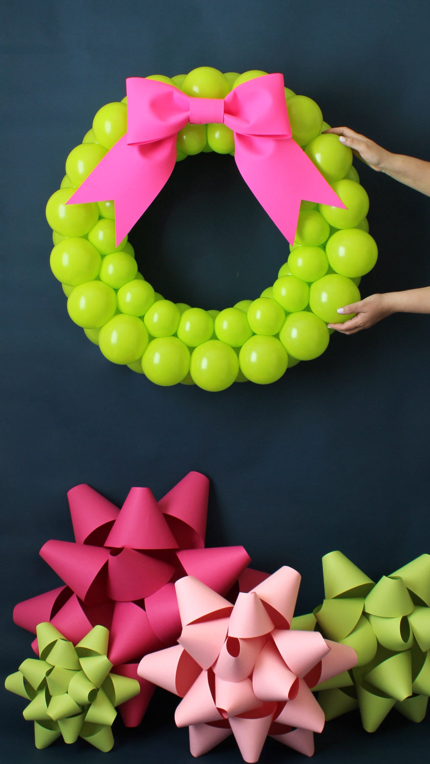 Chartreuse  balloon wreath displayed against a black backdrop, paired with large paper gift bows in pink, red, blush, and green. A vibrant and festive setup perfect for holiday decor, photo booths, and party inspiration.