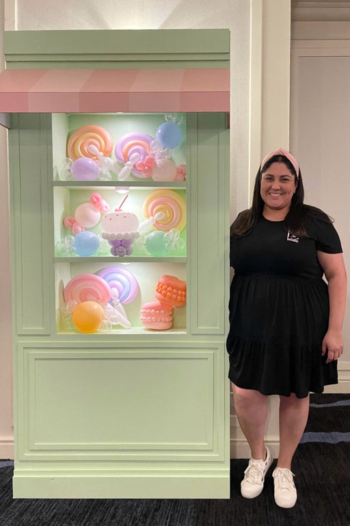 Lilly Jimenez stands beside a pastel green foamboard storefront featuring balloon candy props like swirl lollipops, cupcakes, and macarons inside lighted display shelves. Topped with a pink-and-white striped awning, this DIY prop is part of the Foamboard Storefront Class by The Creative Heart Studio—perfect for event decor, photo backdrops, and party styling.