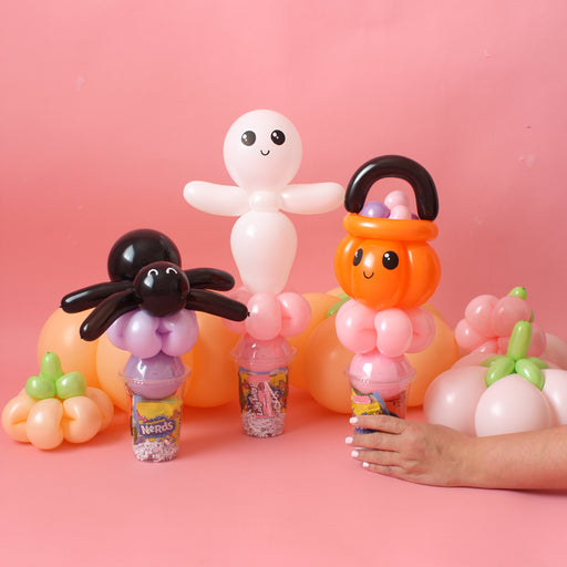 Colorful Halloween balloon candy cup toppers featuring a smiling ghost, a cute pumpkin bucket, and a black spider, each displayed over candy-filled cups with pastel balloon pumpkins in the background. Set against a soft pink backdrop, this playful Halloween balloon craft is perfect for party favors, DIY Halloween treats, or balloon decoration inspiration.