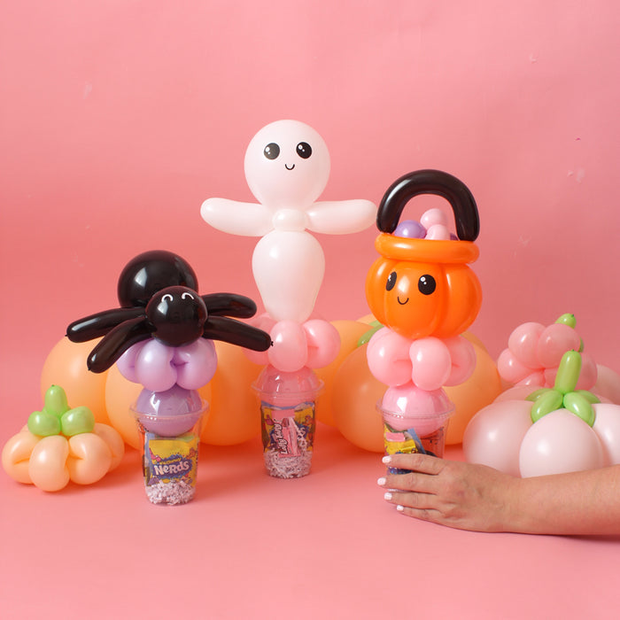 Colorful Halloween balloon candy cup toppers featuring a smiling ghost, a cute pumpkin bucket, and a black spider, each displayed over candy-filled cups with pastel balloon pumpkins in the background. Set against a soft pink backdrop, this playful Halloween balloon craft is perfect for party favors, DIY Halloween treats, or balloon decoration inspiration.