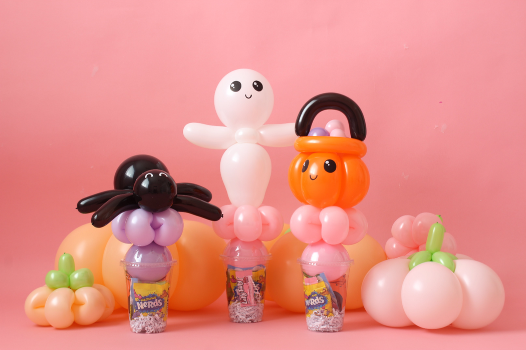 Cute Halloween balloon candy cup toppers featuring a smiling ghost, pumpkin bucket, and spider, each crafted from colorful balloons and placed over Nerds candy cups. Perfect for Halloween parties, trick-or-treat favors, or DIY balloon decor ideas. The pastel pink background and coordinating balloon pumpkins add a fun and festive touch to this Halloween candy craft scene.