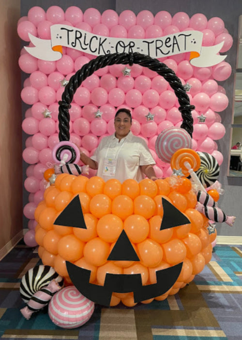 A person poses inside a large balloon pumpkin bucket photo op featuring an oversized jack-o-lantern made of orange balloons with black face details, decorated with balloon candy props and a twisted black handle. The setup includes a pink balloon backdrop with a “Trick or Treat” banner, creating a festive Halloween balloon display for parties, schools, and seasonal events.