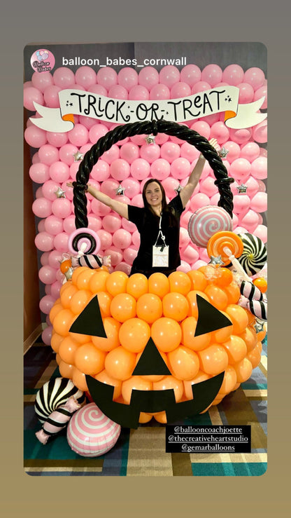 A Halloween balloon pumpkin bucket photo op featuring an oversized orange jack-o-lantern made of balloons, filled with balloon candy decorations and set against a pink balloon wall with a “Trick or Treat” banner. A person poses inside the bucket, showcasing this festive Halloween balloon decor idea for parties, schools, and event backdrops.