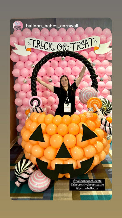 A Halloween balloon pumpkin bucket photo op featuring an oversized orange jack-o-lantern made of balloons, filled with balloon candy decorations and set against a pink balloon wall with a “Trick or Treat” banner. A person poses inside the bucket, showcasing this festive Halloween balloon decor idea for parties, schools, and event backdrops.