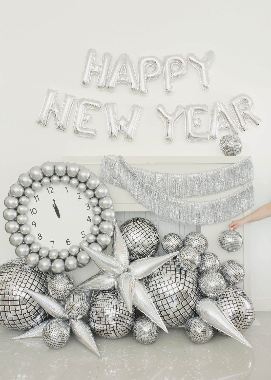 Festive New Year’s Eve balloon decor featuring a large silver balloon clock set to midnight, surrounded by disco ball balloons, chrome starbursts, and a shimmering fringe garland. Silver foil balloons spelling “HAPPY NEW YEAR” complete the celebration scene. This setup is part of Lilly Jimenez’s New Year’s Eve Clock Tutorial—perfect for balloon decorators planning glamorous NYE installations or photo backdrops.