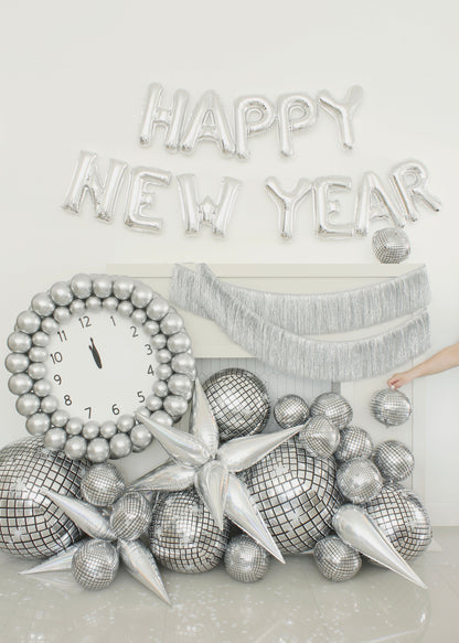 Festive New Year’s Eve balloon decor featuring a large silver balloon clock set to midnight, surrounded by disco ball balloons, chrome starbursts, and a shimmering fringe garland. Silver foil balloons spelling “HAPPY NEW YEAR” complete the celebration scene. This setup is part of Lilly Jimenez’s New Year’s Eve Clock Tutorial—perfect for balloon decorators planning glamorous NYE installations or photo backdrops.
