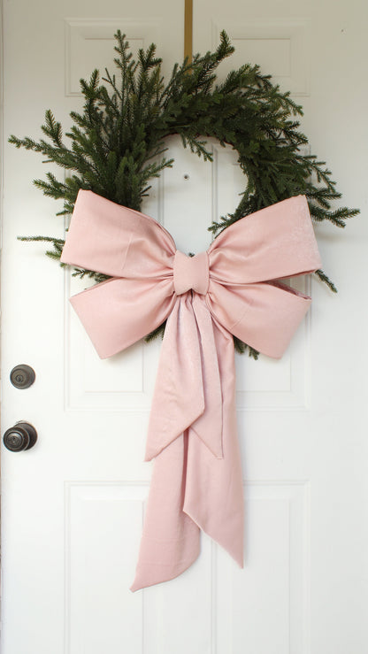 A festive green wreath adorned with an oversized dusty pink fabric bow, hanging on a white door. The bow features elegantly structured loops and long trailing ribbons, adding a soft, elegant touch to the classic holiday decor.