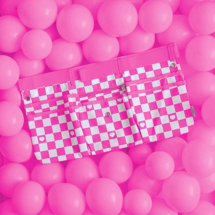 Hot pink and white checkered maker apron with heart detail, designed for balloon artists and event decorators. Features multiple pockets for tools, zip pouch, belt loops, grommets for carabiners, and an adjustable strap. Displayed on a vibrant background of pink balloons, this stylish and functional workbelt is perfect for creative professionals and available in limited quantities for preorder.